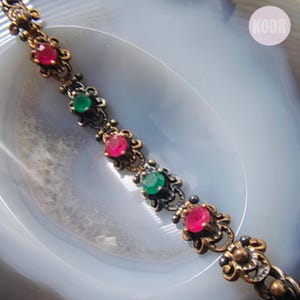 Puede incluir: Un brazalete de estilo antiguo con piedras preciosas rojas y verdes alternas engastadas en metal ornamentado de tono bronce. El diseño del brazalete presenta detalles florales y una delicada cadena.