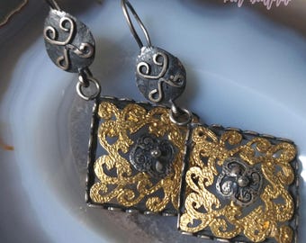 May include: A pair of silver earrings with gold filigree detailing. The earrings feature a square design with intricate floral patterns and a small round accent in the center. The earrings are suspended from a silver hook.