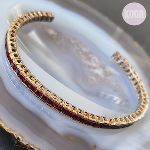 May include: A gold-toned cuff bracelet featuring a continuous row of square-cut deep red stones set in a channel setting. The bracelet has an open design and a metallic finish. A circular logo with the text KOOR is visible in the upper right corner.