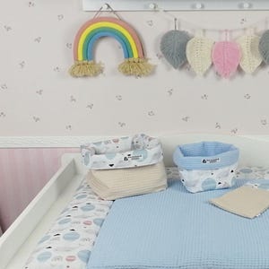 Könnte beinhalten: Ein weißer Wickeltisch mit einer Wickelauflage mit blau-weißem Muster. Es gibt zwei Aufbewahrungskörbe, einen beigen und einen blauen, mit einem weißen und grauen Muster. Die Körbe sind mit gefalteten Handtüchern gefüllt. An der Wand hinter dem Wickeltisch hängen ein Regenbogen-Wandbild und eine Blättergirlande.