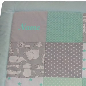 Könnte beinhalten: Eine personalisierte Babydecke mit Patchwork-Design, das verschiedene Tiermotive zeigt, darunter Elefanten, Giraffen und Löwen. Die Decke ist aus einem weichen, hellblauen Stoff gefertigt und hat ein Grau-Weiß-Farbschema. Die Decke ist mit dem Namen "Name" in einer türkisfarbenen Schrift personalisiert.
