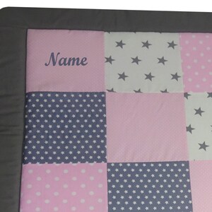Könnte beinhalten: Eine rosa und graue Babydecke mit einem personalisierten Namen, "Name", der in der Mitte gestickt ist. Die Decke besteht aus Patchwork-Quadraten mit verschiedenen Mustern, darunter Sterne, Punkte und kleine Sterne.