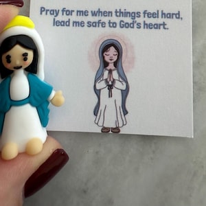 Może przedstawiać: Mała figurka Matki Boskiej, z białą szatą, niebieskim szalem i żółtym nakryciem głowy. Obok kartka z rysunkiem Matki Boskiej i tekstem "Pray for me when things feel hard, lead me safe to God's heart."