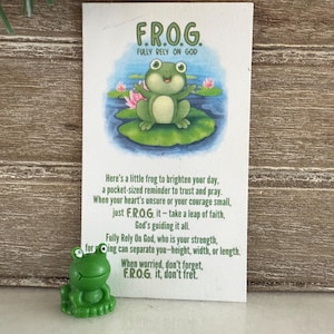 May include: A small, green frog figurine next to a card with the text "F.R.O.G. Fully Rely On God." The card features a cartoon frog on a lily pad. The text on the card offers encouragement and a reminder to trust and pray.
