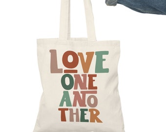 May include: A white canvas tote bag with the words "LOVE ONE ANOTHER" printed in colourful, retro-style letters. The bag has brown straps and is being held by a hand.