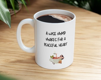 May include: White ceramic coffee mug with a black handle. The mug has black coffee inside and a quote that reads "A wise mind makes for a peaceful heart". There is a cartoon illustration of a woman relaxing in a coffee cup.