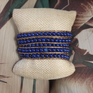 May include: A stack of blue lapis lazuli beaded bracelets with gold accents on a beige fabric background.
