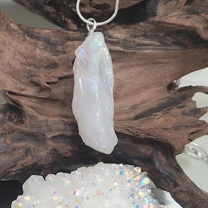 May include: A silver chain necklace with a white iridescent crystal pendant. The crystal is a raw, natural shape with a pointed tip. The necklace is displayed on a piece of driftwood.