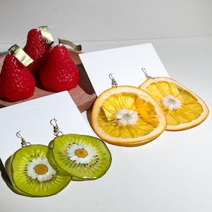 May include: A collection of fruit-themed earrings and strawberry candles. The earrings feature slices of kiwi and orange with a small white flower in the center. The red strawberry candles have gold tags.