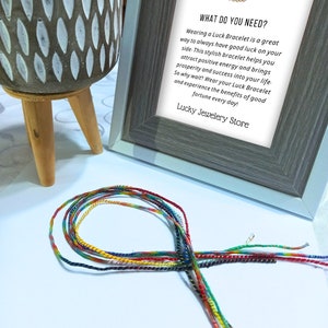 Può includere: Un braccialetto intrecciato multicolore con fili rossi, gialli, verdi e blu. Sullo sfondo una stampa incorniciata con testo sui braccialetti portafortuna. È visibile anche un vaso decorativo con supporto in legno.