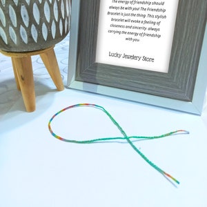 Può includere: Un braccialetto dell'amicizia con un filo color arcobaleno e accenti verdi è appoggiato su una superficie bianca. Una stampa incorniciata dietro recita "Braccialetto dell'amicizia" e include il testo di Lucky Jewelery Store.