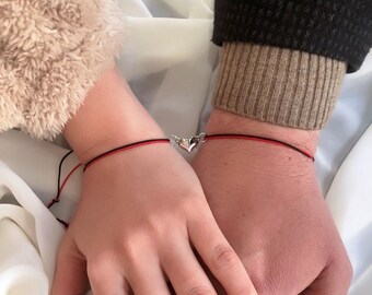 Puede incluir: Dos muñecas adornadas cada una con una pulsera a juego. Las pulseras presentan un cordón negro y rojo y un dije plateado en forma de corazón. Las manos están entrelazadas, lo que sugiere una conexión. Una persona lleva una manga difusa beige, la otra un suéter negro.