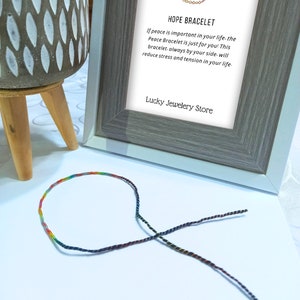 Pode incluir: Uma pulseira da amizade colorida com um design de arco-íris é exibida perto de uma impressão emoldurada. A impressão diz "HOPE BRACELET" e inclui texto sobre paz e redução do estresse. O logotipo da Lucky Jewelery Store está na parte inferior.