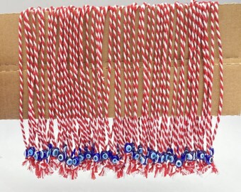 Puede incluir: Una colección de cordones trenzados rojos y blancos, cada uno con una pequeña cuenta de ojo turco azul y blanco en el extremo. Los cordones están dispuestos en una fila ordenada, con las cuentas colgando debajo. Fondo blanco.