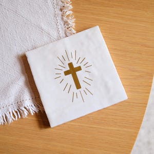 Può includere: Un tovagliolo quadrato bianco con un disegno a croce dorata e linee radiali. Il tovagliolo è su una superficie di legno, accanto a un panno bianco con frange. La croce è centrata e stampata sul tovagliolo.