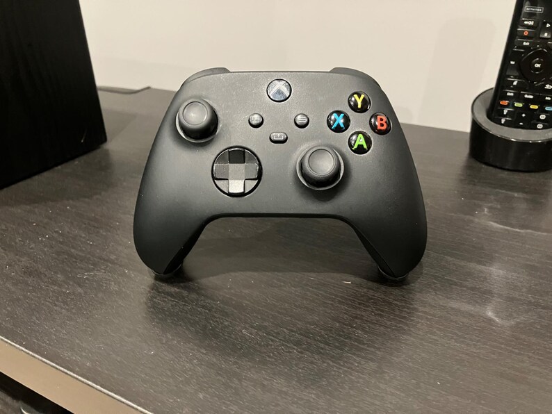 May include: Black Xbox controller with colored buttons, including green A, red B, blue X, and yellow Y. The controller has two analog sticks, a directional pad, and several other buttons. The Xbox logo is visible at the top.