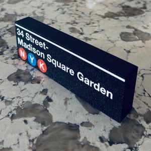 May include: A rectangular black block sign featuring white text that reads 34 Street-Madison Square Garden. Below the text are three circular icons in red, blue, and red, containing the white letters N, Y, and K respectively, on a textured surface.