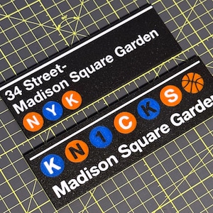 May include: Two rectangular black signs with white text and colorful circular accents. One sign reads 34 Street-Madison Square Garden with NYK in circles. The other reads KNICKS Madison Square Garden with a basketball icon and circular letters.