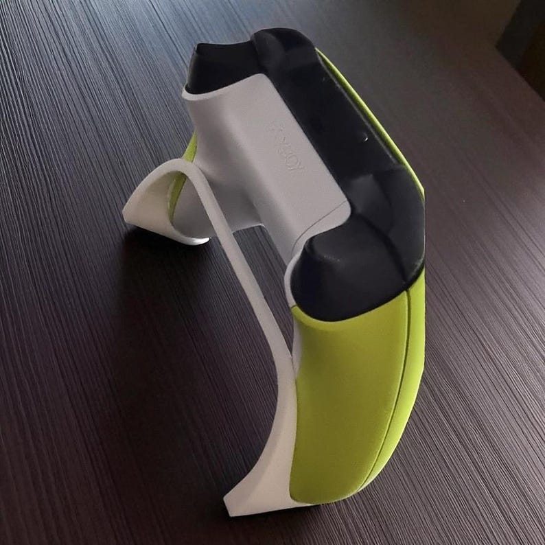 May include: A gaming controller stand in white, black, and lime green. The stand is designed to hold a gaming controller. The word "XBOX" is visible on the controller. The stand is on a dark brown surface.