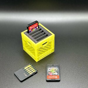 May include: A yellow 3D printed game cartridge holder with slots for multiple cartridges. A Nintendo Switch game cartridge is in the top slot. Two additional game cartridges are on the table next to the holder.