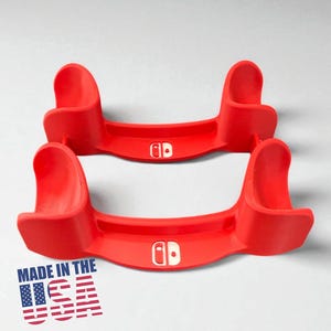 May include: Two bright red game console stands. Each stand has a curved design with a white logo. The stands are labeled "MADE IN THE USA" with a graphic of the American flag.
