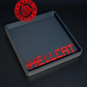 May include: A dark gray rectangular tray with a red "HELLCAT" logo. The tray has a raised edge and a "MADE IN AMERICA" emblem. The tray is likely for automotive parts or accessories.
