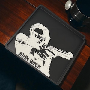 May include: Black square tray featuring a white graphic of a man holding a gun, with the text "JOHN WICK" in white. The tray has a dark gray border.
