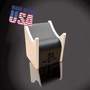 May include: A white and black PlayStation controller stand with the PlayStation logo on the front. The stand is made of 3D printed plastic and has a curved design. The stand is made in the USA.