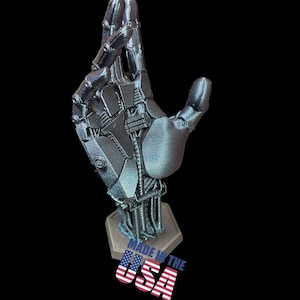 May include: A silver metallic 3D printed robotic hand sculpture with a textured surface. The hand is posed with the index and middle fingers extended and the other fingers curled. The sculpture is mounted on a hexagonal base with the words "Made in the USA" printed on the base.