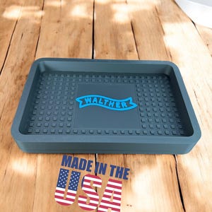 May include: A gray rubber doormat with a raised textured surface and a blue banner with the word "WALTHER" printed on it. The doormat is on a wooden surface and has the words "MADE IN THE USA" printed below it.