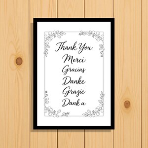 May include: A rectangular black frame containing a white print with a black line-art eucalyptus border. The text reads: Thank You, Merci, Gracias, Danke, Grazie, Dank u. The design features elegant black cursive lettering centered within the frame.