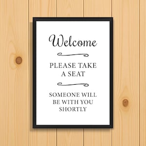 May include: A framed sign with the word "Welcome" in elegant script, followed by "PLEASE TAKE A SEAT" and "SOMEONE WILL BE WITH YOU SHORTLY" in a black frame. The sign is mounted on a light brown wooden background.