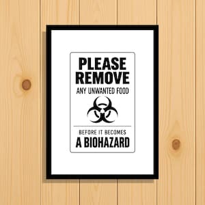 Könnte beinhalten: Ein rechteckiger schwarzer Rahmen mit einem weißen Schild, das den schwarzen Text trägt: PLEASE REMOVE ANY UNWANTED FOOD BEFORE IT BECOMES A BIOHAZARD. Das Schild zeigt ein zentrales schwarzes Biohazard-Symbol zwischen den Textzeilen.