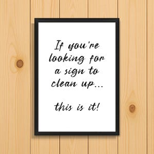 Puede incluir: Un cartel enmarcado con el texto "If you're looking for a sign to clean up... this is it!" en escritura negra sobre un fondo blanco. El marco es negro y el fondo es de vetas de madera clara.