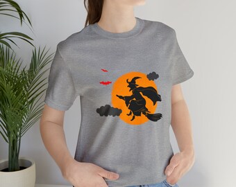Puede incluir: Una camiseta gris jaspeada con una silueta negra de una bruja volando en una escoba frente a una luna naranja brillante. Los murciélagos vuelan en el cielo sobre la luna.