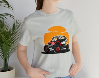 May include: A white t-shirt featuring a graphic of a red and black off-road vehicle driving into a sunset. The vehicle has black wheels and a roll cage.