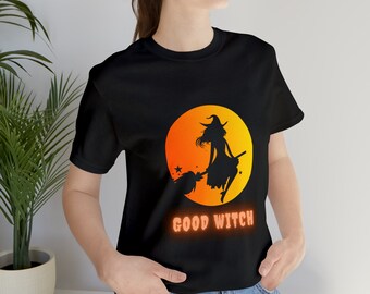 Puede incluir: Camiseta negra con un gráfico de una bruja volando en una escoba frente a una luna de puesta de sol. El texto "GOOD WITCH" está impreso debajo del gráfico.