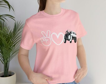 May include: A pink t-shirt with a white peace sign, heart, and a black and white jeep graphic. The text "Peace Love Jeep" is printed on the shirt.