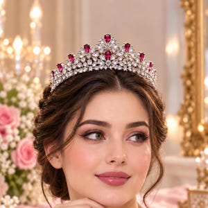 May include: A silver-toned tiara featuring a series of teardrop-shaped red stones arranged along the top edge, interspersed with numerous clear, faceted crystals set in a dense, intricate metal framework that forms a pointed, crown-like structure.