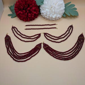 May include: A set of five burgundy crystal beaded necklaces with gold clasps. The necklaces are arranged in a semi-circle shape on a light beige surface.