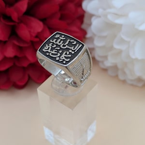 May include: Silver ring with a square face bearing Arabic calligraphy. The ring has a textured band and is displayed on a clear stand against a backdrop of red and white flowers. The ring's design is detailed and ornate.