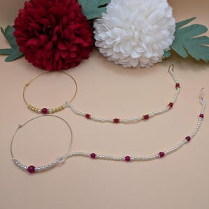 May include: Two gold nose rings with white pearl and red bead accents. The rings are on a light beige surface with white and red flowers.