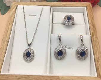 May include: A silver jewellery set featuring a blue oval gemstone pendant, matching earrings, and a ring. The jewellery is presented in a wooden box with white velvet lining.