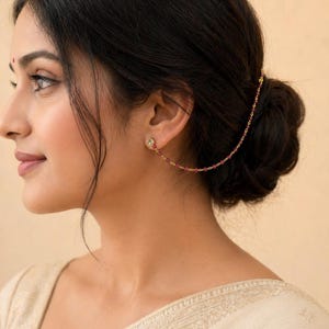 May include: A gold-toned earring with a small, clear gem and a chain of small, red beads. The chain extends from the earring to the hair, creating a delicate, elegant look. The jewelry is worn on the ear.