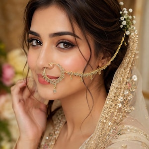 May include: A gold nose ring featuring intricate metalwork and small clear stones, connected by a delicate gold chain to a hair accessory adorned with tiny white flowers. The nose ring has a circular frame with a dangling central stone element.