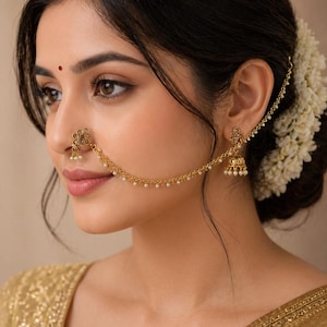 May include: A gold-coloured nose ring and earring set with small pearl details. The nose ring has a floral pattern, linked by a chain of tiny pearls to a matching earring with a bell-shaped charm. The jewellery is worn on a person.