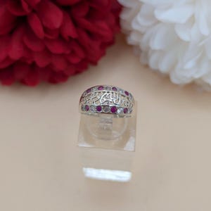 Könnte beinhalten: Ein silberner Ring mit einem aufwendigen Design mit roten und klaren Edelsteinen. Der Ring hat ein verziertes Muster mit arabischer Schrift. Der Ring wird auf einem transparenten Ständer präsentiert.