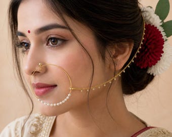 May include: A gold-colored nose ring with a pearl-lined hoop and a delicate chain with small pearl accents. The jewelry is worn by a person with dark hair, a red bindi, and a flower hair accessory.