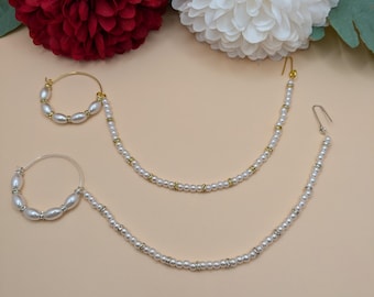 May include: Two gold nose rings with white pearl beads and a gold chain. The nose rings are on a light brown surface with red and white flowers in the background.