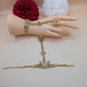 May include: A gold hand chain bracelet with intricate detailing. The bracelet features square and round decorative elements with red, green, and white accents. The ring is connected to the bracelet by a delicate chain.